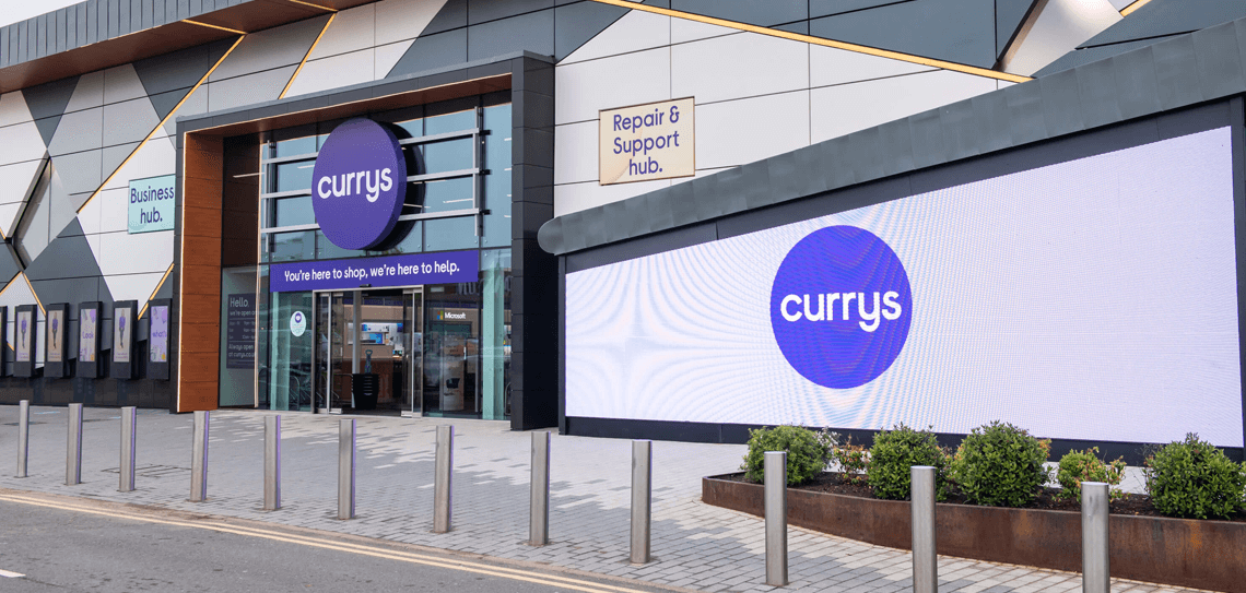 Front of modern Currys store with large advertising screen to right and shrubs bottom right with parking bollards across bottom of image.